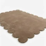 Angled perspective view of the Amber Glint rectangular wool rug displaying the warm tan color and soft scalloped border.