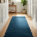 Perspective lifestyle view of the Aqualene runner rug placed on a warm wooden floor in a sunlit hallway with crisp white wainscoting, featuring a solid deep teal blue color and a continuous contoured scalloped edge.