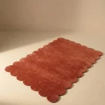 Angled perspective view of the Clay Ember rectangular wool rug displaying the warm terracotta color and soft scalloped border.