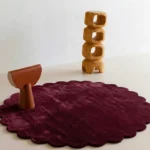 Crimson Aura round wool rug with scalloped edges displayed in a modern minimalist room setting with wooden abstract decor.