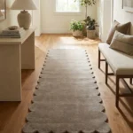 Perspective lifestyle view of the Driftstone runner rug placed on a warm wooden floor in a brightly lit hallway with a console table and bench, featuring a solid light beige color and a continuous contoured scalloped edge.