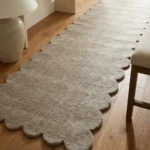 Close-up angled view of the driftstone runner rug placed on a warm wooden floor next to a console table, highlighting the dense, plush hand-tufted New Zealand wool and viscose silk texture and the gracefully contoured deep gray scalloped border.