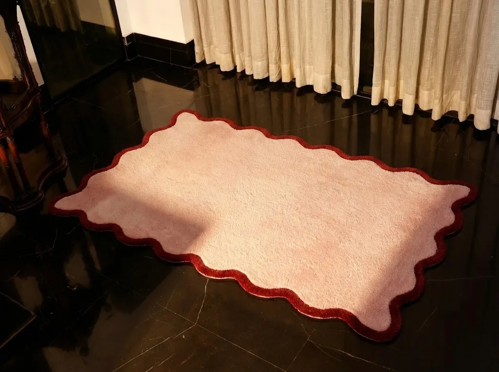 A completed hand-tufted scalloped rug in soft pink with a dark red border, lying on a polished black floor.