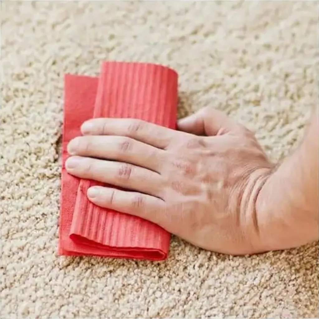 Hands using a clean red cloth to gently blot a stain on a wool rug demonstrating proper spot cleaning technique.
