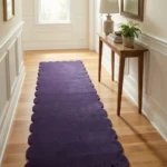 Perspective lifestyle view of the Midnight Serenade runner rug placed on a warm wooden floor in a brightly lit hallway, featuring a solid deep purple color and a continuous contoured scalloped edge extending down the corridor.