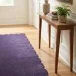 Close-up angled view of the Midnight Serenade runner rug placed on a warm wooden floor next to a console table, highlighting the dense, plush hand-tufted New Zealand wool and viscose silk texture and the gracefully contoured deep purple scalloped border.