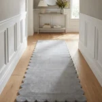 Perspective lifestyle view of the Moonhaze runner rug placed on a warm wooden floor in a sunlit hallway with crisp white wainscoting, featuring a solid light grey color and a continuous contoured scalloped edge.