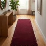 Perspective lifestyle view of the Ruby Ember runner rug placed on a warm wooden floor in a brightly lit hallway, featuring a solid deep burgundy red color and a continuous contoured scalloped edge.