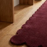 Close-up angled view of the Ruby Ember runner rug on a wooden floor, highlighting the dense, plush hand-tufted New Zealand wool and viscose silk texture and the gracefully contoured deep burgundy red scalloped border.