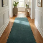 Perspective lifestyle view of the Tidefall runner rug placed on a warm wooden floor in a brightly lit hallway decorated with natural greenery, featuring a solid muted teal blue color and a continuous contoured scalloped edge.