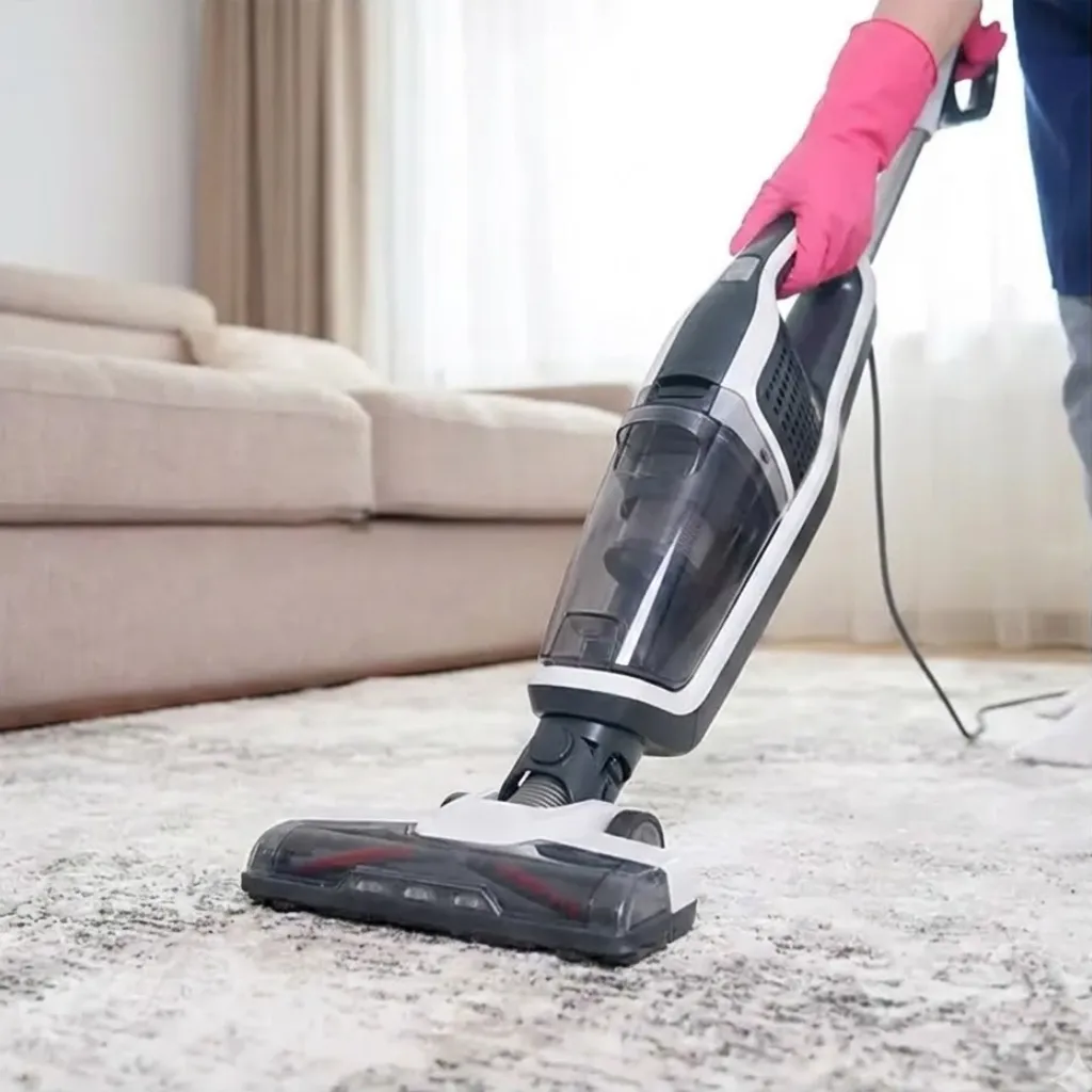 Person using a modern stick vacuum cleaner on a light colored area rug to demonstrate long term care and maintenance.