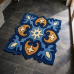 High angle view of the Azure Heirloom square rug placed on a dark tiled floor, featuring an ornate, traditional-inspired floral motif in vibrant light blue, deep navy, gold, and cream, finished with a complex contoured scalloped edge.