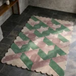 Perspective view of the Blushed Olive rug placed on a dark tiled floor, showcasing the bold 3D geometric lattice design and the structured cream scalloped border illuminated by natural sunlight.