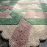 Close-up angled view of the Blushed Olive rug, highlighting the dense hand-tufted New Zealand wool texture, the deeply carved lines defining the interlocking green, pink, and cream geometric shapes, and the intricately contoured scalloped edge.
