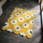 Perspective view of the Butter Pop rug placed on a dark tiled floor, featuring a bright yellow background adorned with a repeating retro pattern of white flowers with black centers, finished with a petal-contoured scalloped edge.
