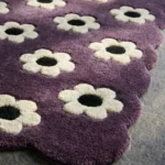 Close-up low angle view of the Daisy Night rug, showcasing the plush hand-tufted wool texture, the deeply carved white daisy flowers with black centers, and the intricately contoured purple scalloped border.