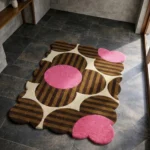 Perspective view of the Dusky Bloom rug placed on a dark tiled floor, featuring a retro oversized floral pattern with brown and tan striped petals, bright pink circular centers, and cream accents, finished with a petal-contoured scalloped edge.