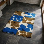 Perspective view of the Earthen Dusk rug placed on a dark tiled floor, featuring a retro oversized floral pattern in deep blue and earthy brown on a cream background, finished with a petal-contoured scalloped edge.