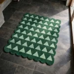 Perspective view of the Emerald Steps rug placed on a dark tiled floor, showcasing the bold repeating geometric pattern of light green triangles on a solid dark green background, finished with a structured scalloped border illuminated by natural sunlight.