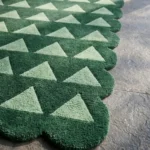 Close-up angled view of the Emerald Steps rug, highlighting the plush hand-tufted New Zealand wool and viscose silk texture, the deeply carved lines defining the light green geometric triangles against the dark green background, and the intricately contoured scalloped edge.