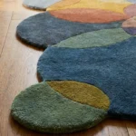 Close-up angled view of the Eucalyptus Veil runner rug placed on a warm wooden floor, highlighting the dense, plush hand-tufted New Zealand wool and viscose silk texture. The design features a playful geometric pattern of overlapping circles in a muted, earthy palette of eucalyptus green, dusty blue, mustard yellow, slate grey, and burnt orange, which naturally form a dynamic, wavy contoured scalloped edge