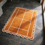 Perspective view of the Hearth Grid rug placed on a dark tiled floor, featuring a warm rust orange background with a minimalist white windowpane grid pattern, finished with a decorative white scalloped edge.