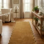 Perspective lifestyle view of the Honeystone runner rug placed on a warm wooden floor in a brightly lit living area, featuring a solid mustard yellow color and a continuous contoured scalloped edge.