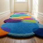 Perspective lifestyle view of the Indigo Carnival runner rug placed on a warm wooden floor in a brightly lit hallway, featuring a playful geometric pattern of overlapping circles in vibrant, bold hues of royal blue, deep purple, bright orange, yellow, green, and pink. The intersecting circular shapes naturally form a dynamic, wavy scalloped edge along the length of the plush, hand-tufted New Zealand wool and viscose silk runner.