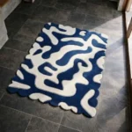 Perspective view of the Inked Dyed rug placed on a dark tiled floor, showcasing the fluid, organic abstract design and the dynamically sculpted scalloped border illuminated by natural sunlight.