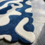 Close-up angled view of the Inked Dyed rug, highlighting the plush hand-tufted New Zealand wool and viscose silk texture, the deeply carved lines defining the abstract blue and cream shapes, and the intricately contoured scalloped edge.