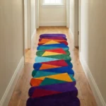 Perspective lifestyle view of the Jewel Shards runner rug placed on a warm wooden floor in a brightly lit hallway, featuring a vibrant geometric patchwork pattern of intersecting triangles in rich jewel tones of red, orange, yellow, green, blue, and purple, naturally forming a wavy contoured scalloped edge.