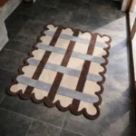 High angle view of the Linen Blend rug featuring an overlapping woven lattice pattern in earthy brown, light blue, and cream tones, finished with a brown scalloped edge.