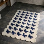Perspective view of the Midnight Surge rug placed on a dark tiled floor, showcasing the bold repeating 3D geometric cross design and the structured cream scalloped border illuminated by natural sunlight.