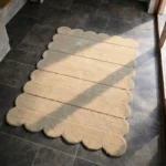 Perspective view of the Milky Ridges rug placed on a dark tiled floor, featuring a solid cream color with horizontally carved ribbed lines and a continuous scalloped edge.
