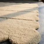 Close-up detail of the Milky Ridges rug illuminated by sunlight, showcasing the dense hand-tufted wool texture, the deeply embossed horizontal lines, and the intricately carved cream scalloped border.