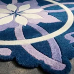 Close-up low angle view of the Moon Blossom rug, showcasing the dense hand-tufted wool texture, the deeply carved lines defining the blue, lavender, and cream interlocking floral pattern, and the intricately contoured scalloped edge.