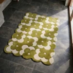 Perspective view of the Moss Parade rug placed on a dark tiled floor, featuring a repeating retro floral pattern in earthy moss green and cream, finished with a petal-contoured scalloped edge.