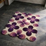 Perspective view of the Mulberry Petal rug placed on a dark tiled floor, featuring a retro pattern of large four-petal flowers in shades of deep purple and soft pink on a cream background, finished with a petal-contoured scalloped edge.