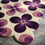 Close-up low angle view of the Mulberry Petal rug, showcasing the plush hand-tufted wool texture, the deeply carved lines separating the deep purple, light purple, and pink flower petals, and the intricately contoured scalloped border.