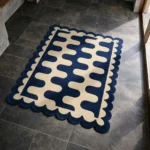 High angle view of the Obsidian Grid rug placed on a dark tiled floor, featuring a bold interlocking wavy pattern in deep blue and cream, finished with a decorative scalloped edge.