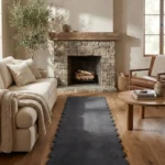 Perspective lifestyle view of the Obsira runner rug placed on a warm wooden floor in a cozy living room with a stone fireplace, featuring a solid dark grey color and a continuous contoured scalloped edge.