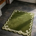 High angle view of the Olive Reverie rug featuring a lush olive green wool background with a decorative cream ribbon loop border and elegant scalloped edges.
