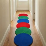 Perspective lifestyle view of the Pastel Ring Stack runner rug placed on a warm wooden floor in a brightly lit hallway, showcasing the dynamic, repeating pattern of overlapping red, green, and blue circles and the playful wavy scalloped silhouette extending down the corridor.