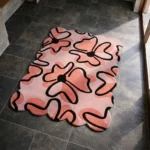 Perspective view of the Peachy Sketch rug placed on a dark tiled floor, featuring a soft peach and pink background adorned with large, abstract floral motifs outlined in thick black lines, finished with a deeply contoured scalloped edge.