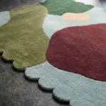 Close-up angled view of the Pebble Parade rug, highlighting the dense hand-tufted wool texture, the deeply carved lines separating the colorful organic abstract shapes, and the intricately contoured light blue scalloped edge.