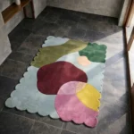 Perspective view of the Pebble Parade rug placed on a dark tiled floor, showcasing the bold abstract pebble design and the structured light blue scalloped border illuminated by natural sunlight.