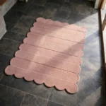 Perspective view of the Petal Layers rug placed on a dark tiled floor, featuring a solid soft pink color with horizontally carved ribbed lines and a continuous scalloped edge.