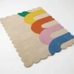 Angled perspective view of the Rainbow Arcana hand tufted rug placed on a white floor showing the playful multicolor pattern and decorative wavy scalloped edges.