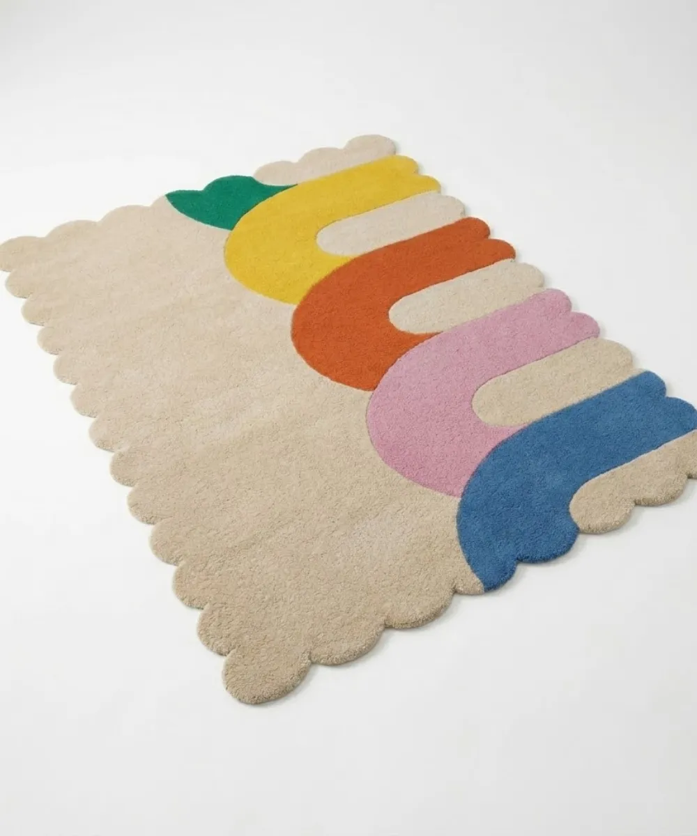 Angled perspective view of the Rainbow Arcana hand tufted rug placed on a white floor showing the playful multicolor pattern and decorative wavy scalloped edges.