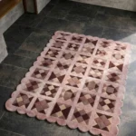 Perspective view of the Rose Matrix rug placed on a dark tiled floor, showcasing the bold geometric patchwork design and the structured pink scalloped border illuminated by natural sunlight.